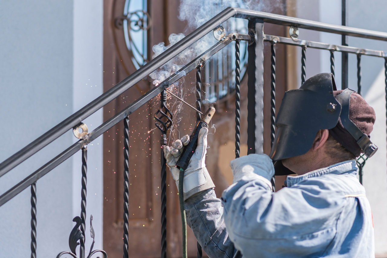 a welder wearing protective gloves and a welding mask repairs a metal angle bar of a fence